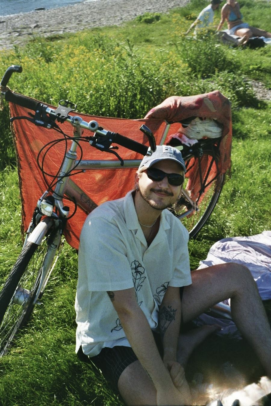 Me in a cap and sunglasses, sitting in the grass next to my bike